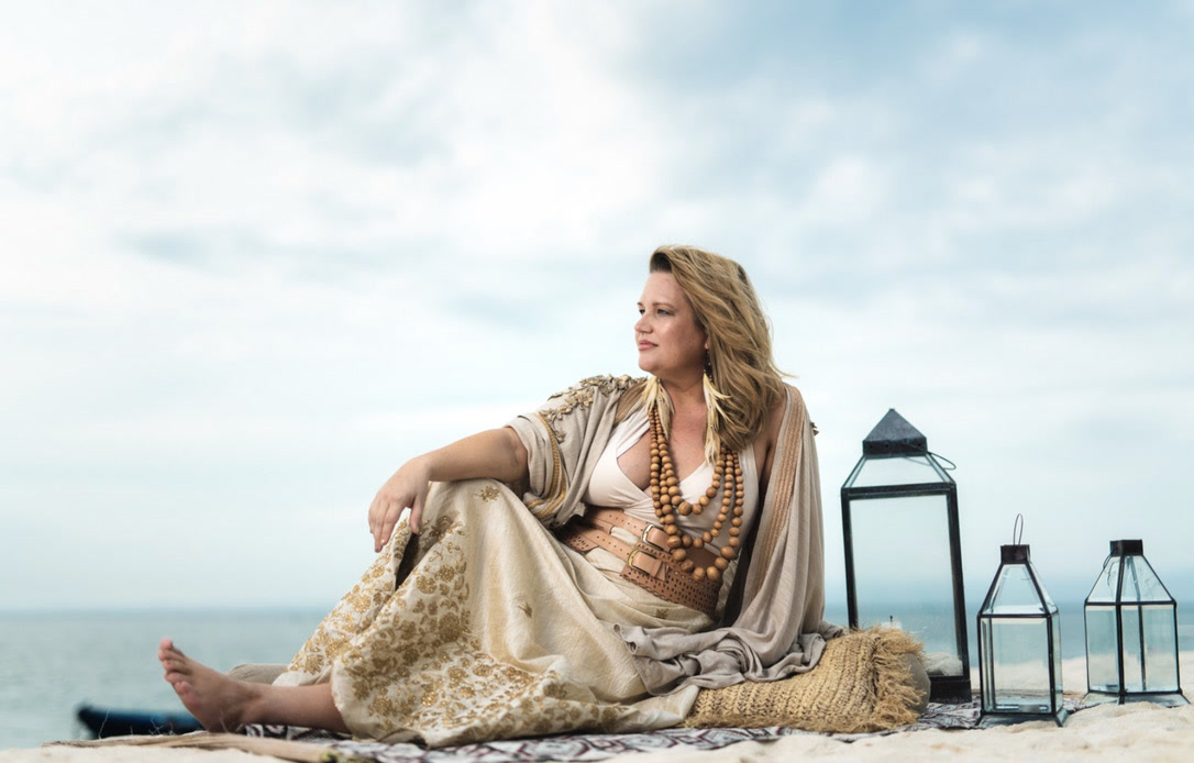 Heather seated barefoot on a woven rug on the sand, in an embroidered skirt, wooden bead necklaces and a leather belt, gazing out to sea beside three glass lanterns.