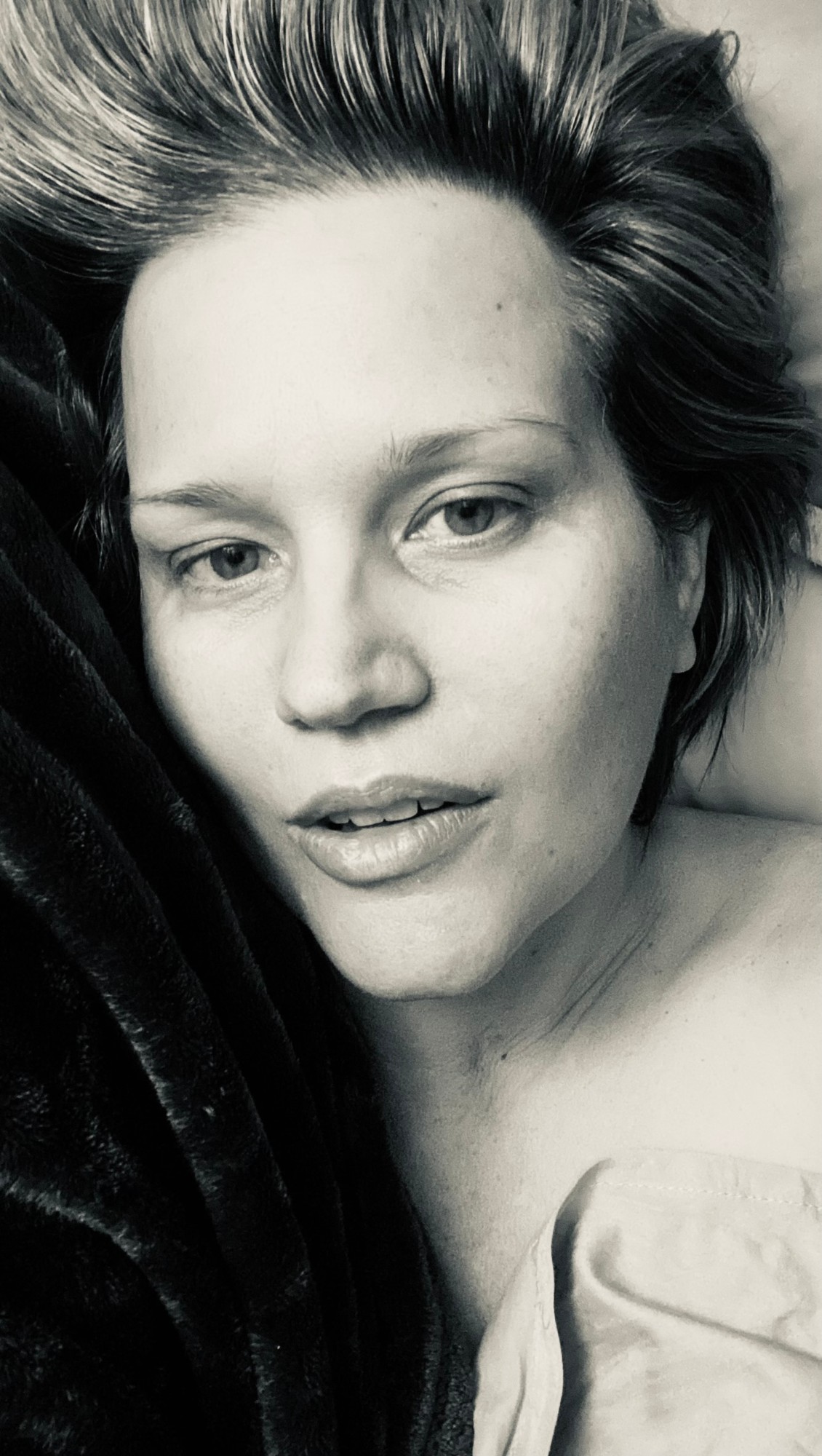 An intimate black and white self portrait of Heather lying back on a pillow, bare shouldered, short hair swept up, looking softly into the lens.