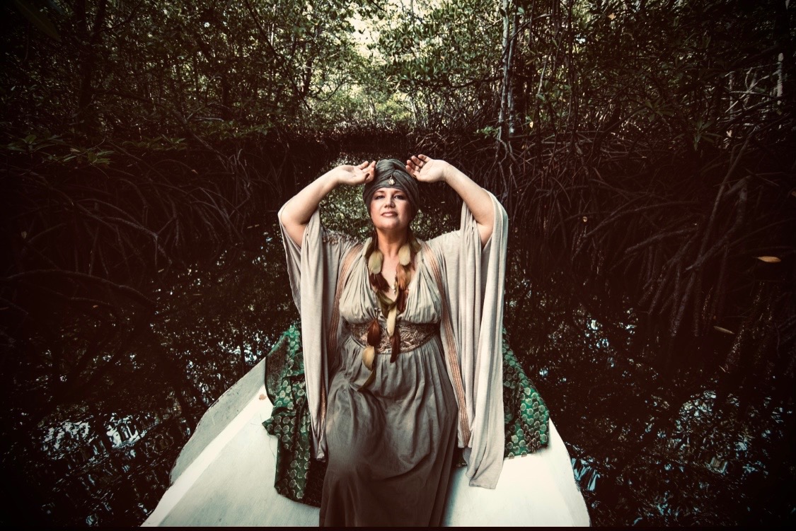 Heather Brand stands at the bow of a white boat in a mangrove channel, arms raised above her turban, in a long flowing dress with feathers braided through her hair.