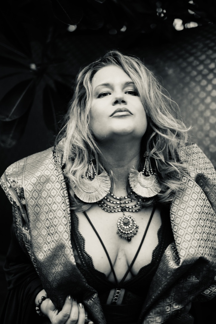 Black and white portrait of Heather, chin lifted, looking straight into the lens in a brocade wrap with fan earrings and layered necklaces.