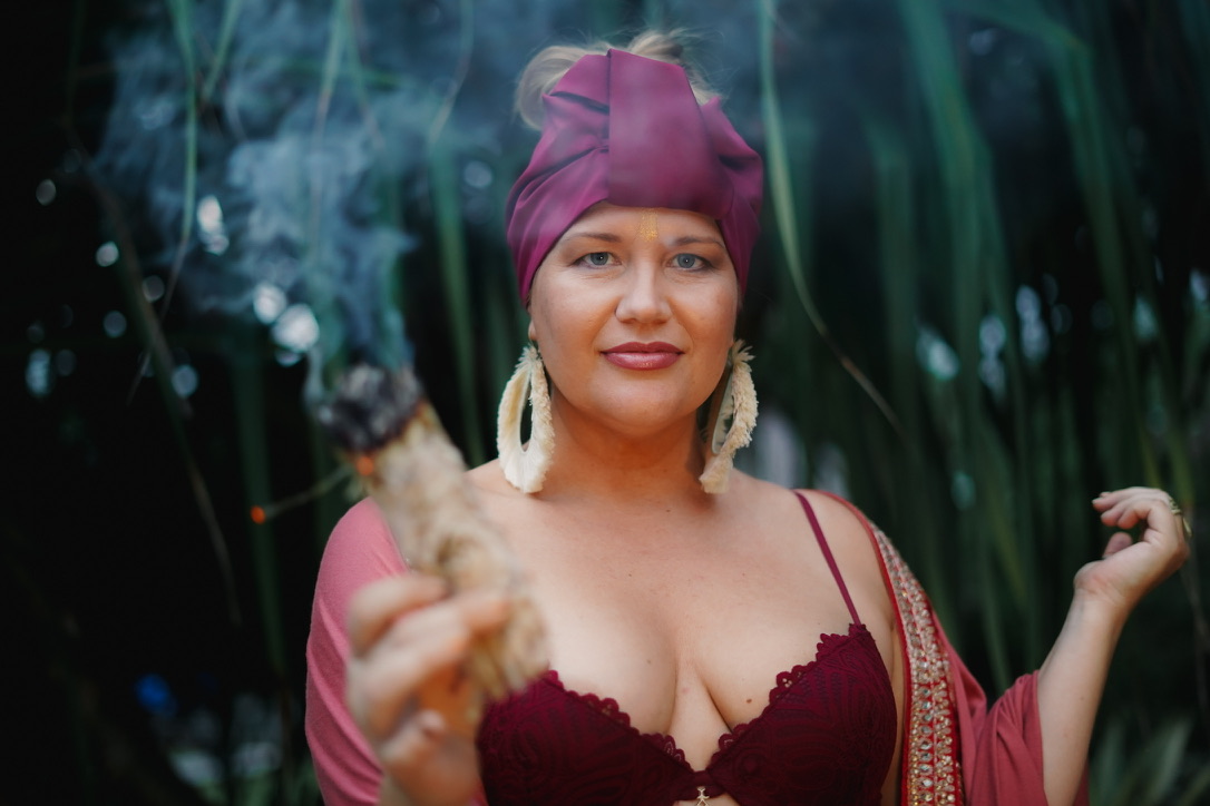 Heather in a magenta turban and deep red lace, holding a smoking bundle of sage toward the camera in front of tall grasses.