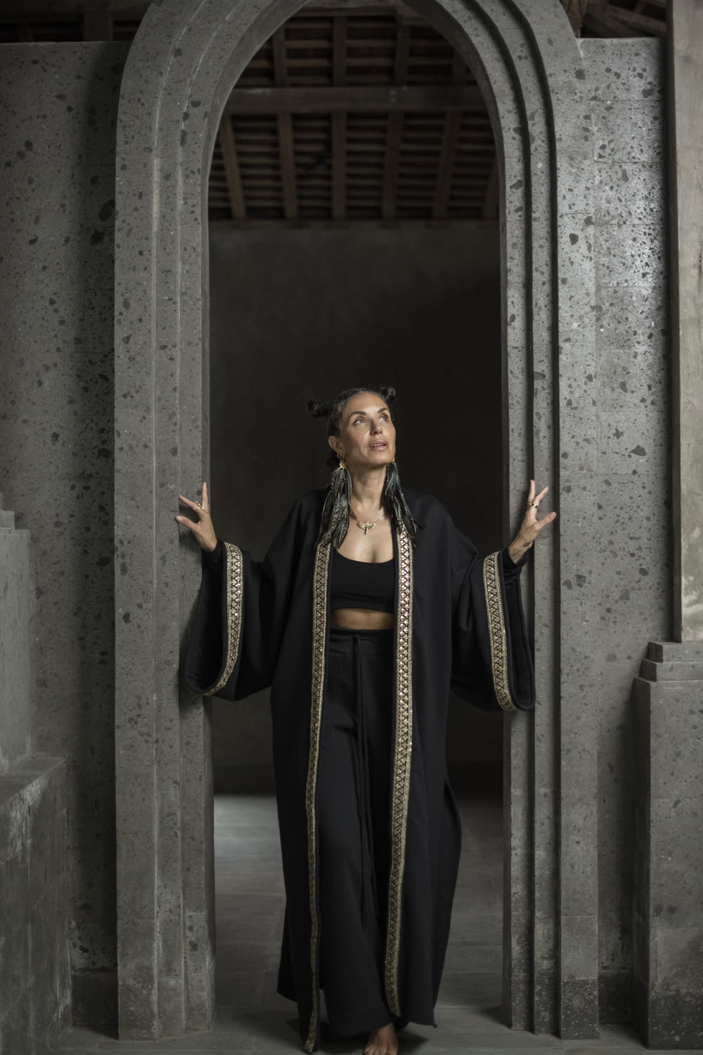 A model in a black fleece coat with gold borders stands in a stone archway, hands raised to the walls.