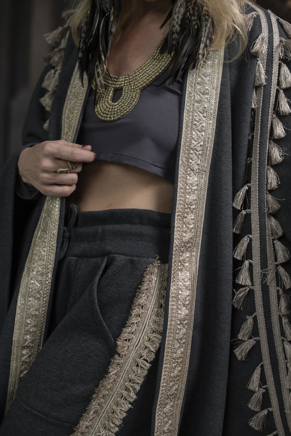 Close detail of a charcoal fleece cape with gold trim and tassels over a cropped top and black trousers.
