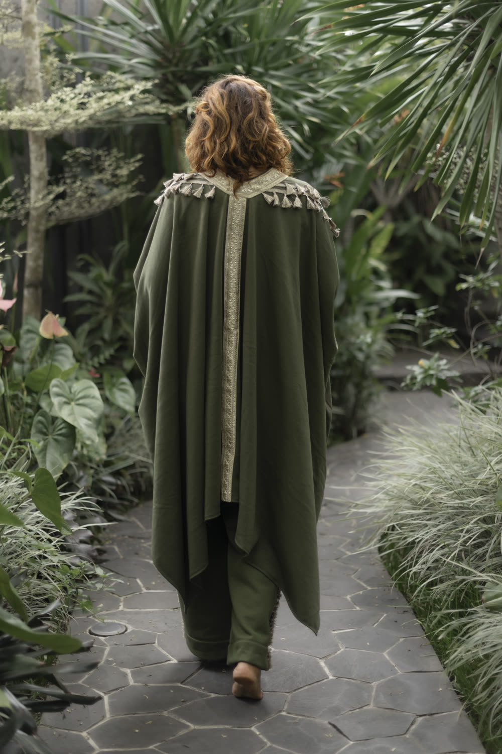 A woman seen from behind walking a garden path in an olive fleece cape with a gold collar.