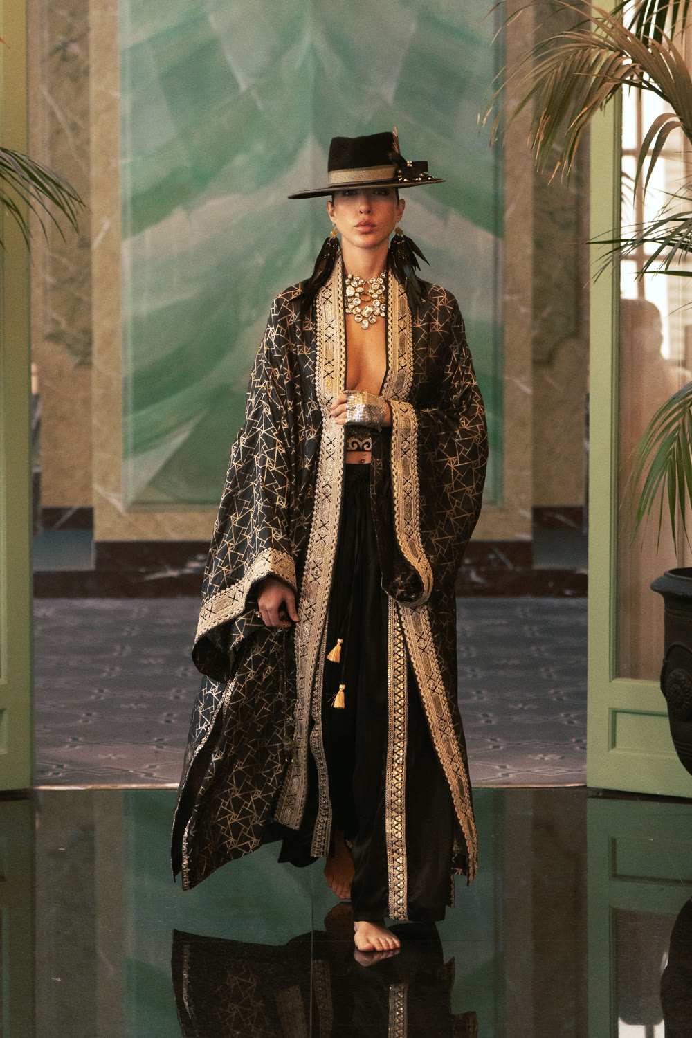 A model in a wide black hat and a long black and gold brocade coat on the runway.