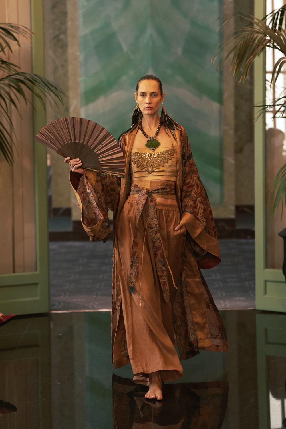 A model holding a fan in a long amber brocade robe on the runway.