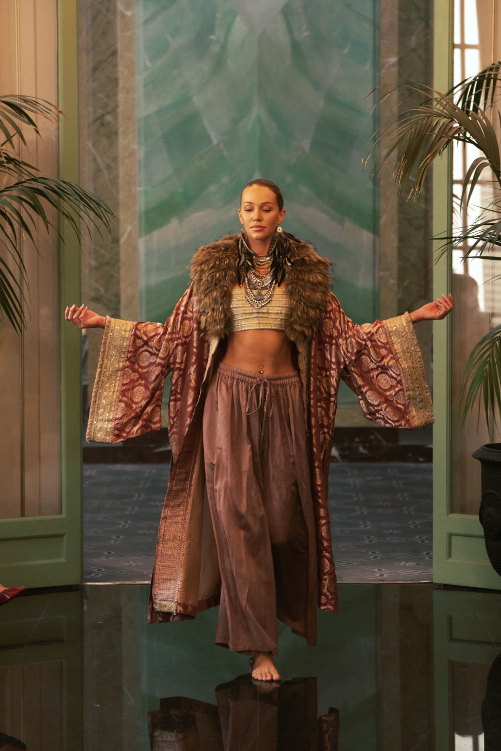 A model with arms open in a rust and gold kimono, embroidered bralette and wide pant on the runway.
