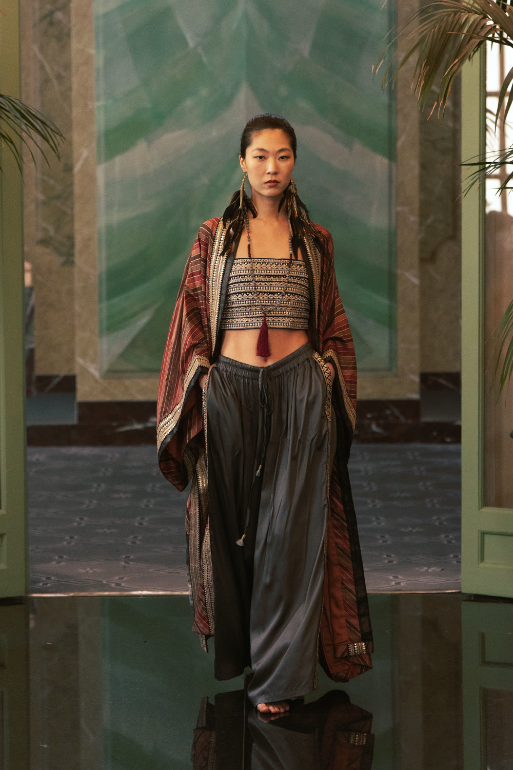 A model in a striped red and gold kimono, embroidered bralette with a tassel and grey wide pant on the runway.
