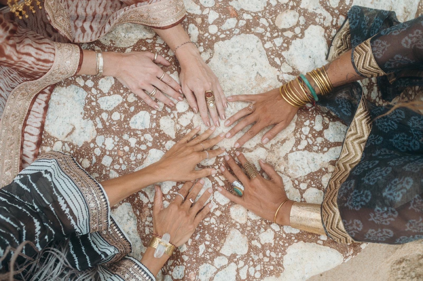 Seen from above, the hands of five women in bangles, rings and cuffs meeting in a circle on a stone table.