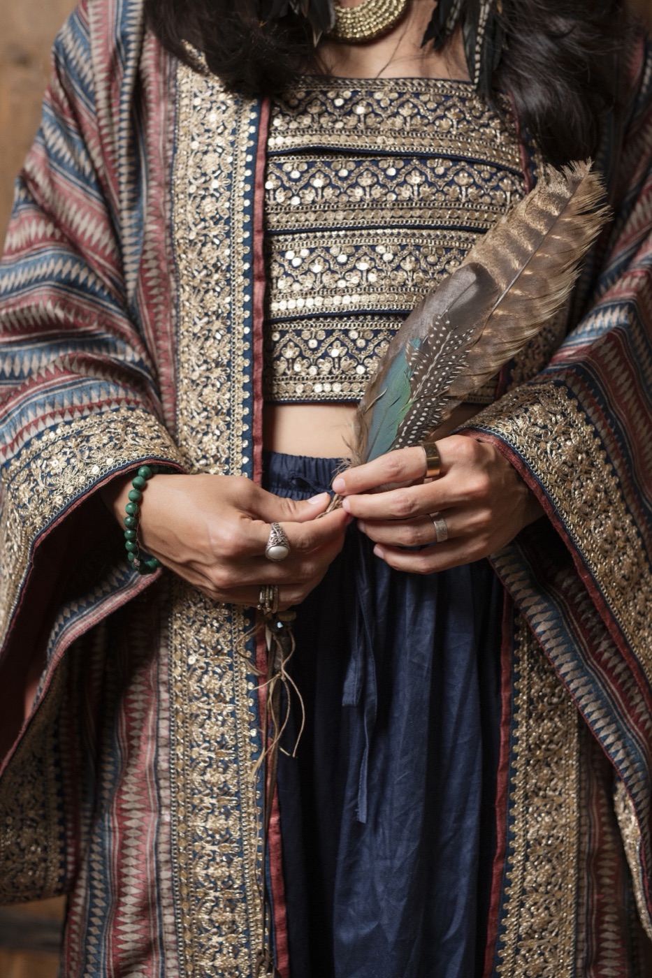 Hands with rings holding a feather against a brocade kimono over an embroidered bralette and indigo skirt.