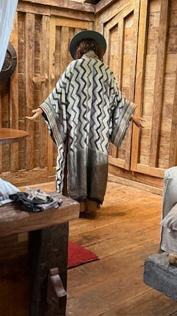 A woman in a hat seen from behind in a black and cream zigzag robe, arms open.