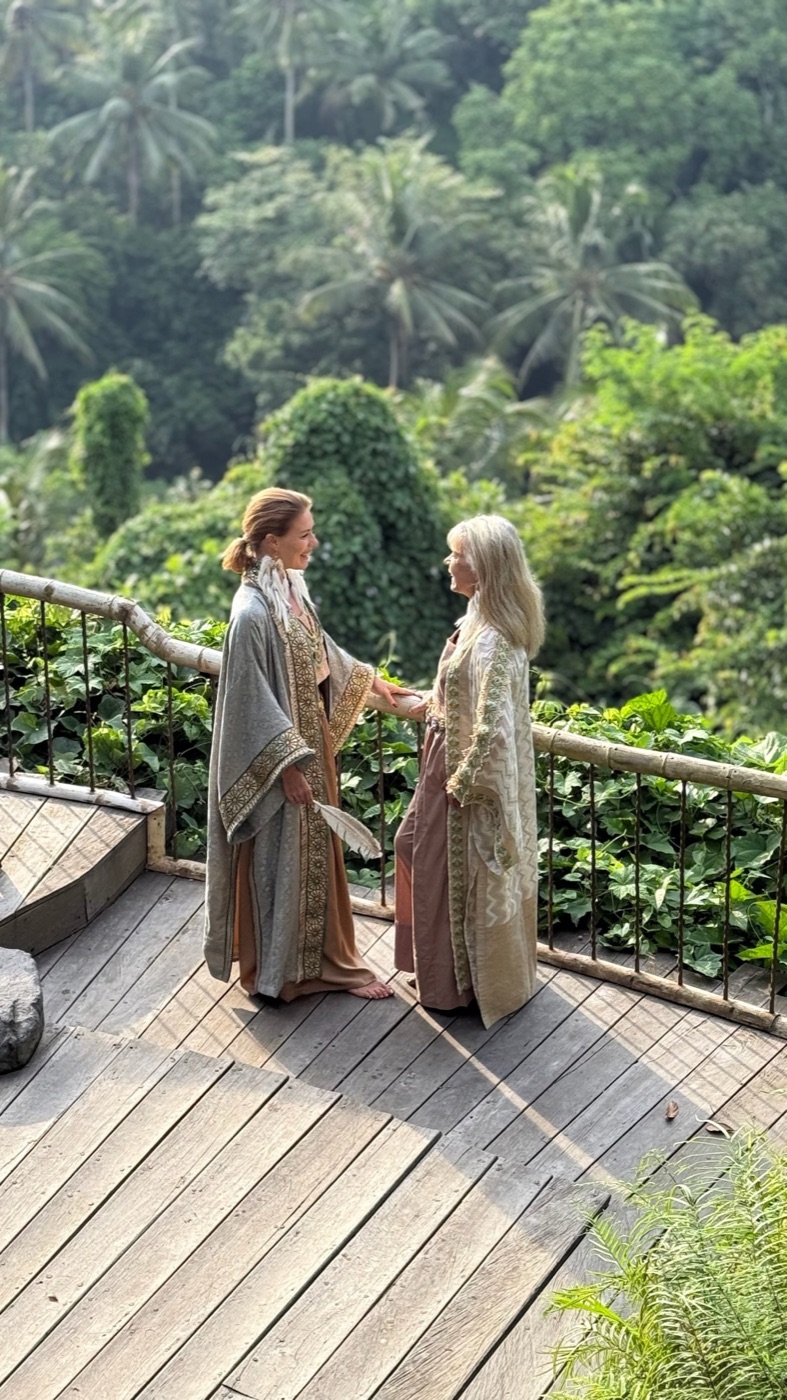 Two women in long robes holding hands on a wooden deck above the Bali jungle and rice fields.