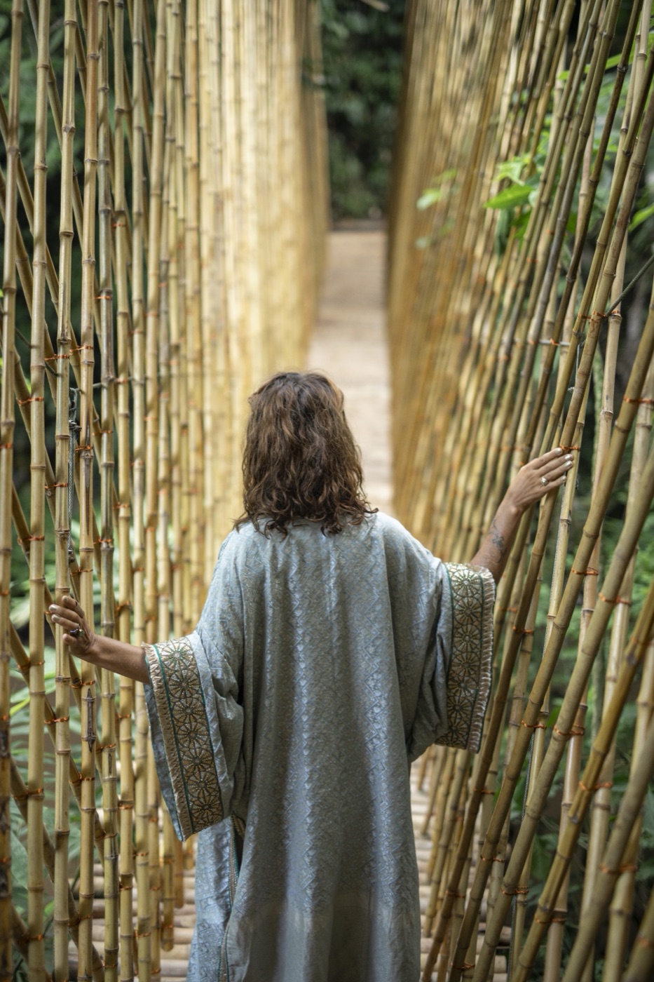 A woman seen from behind in a sage silk robe, arms open between walls of bamboo.