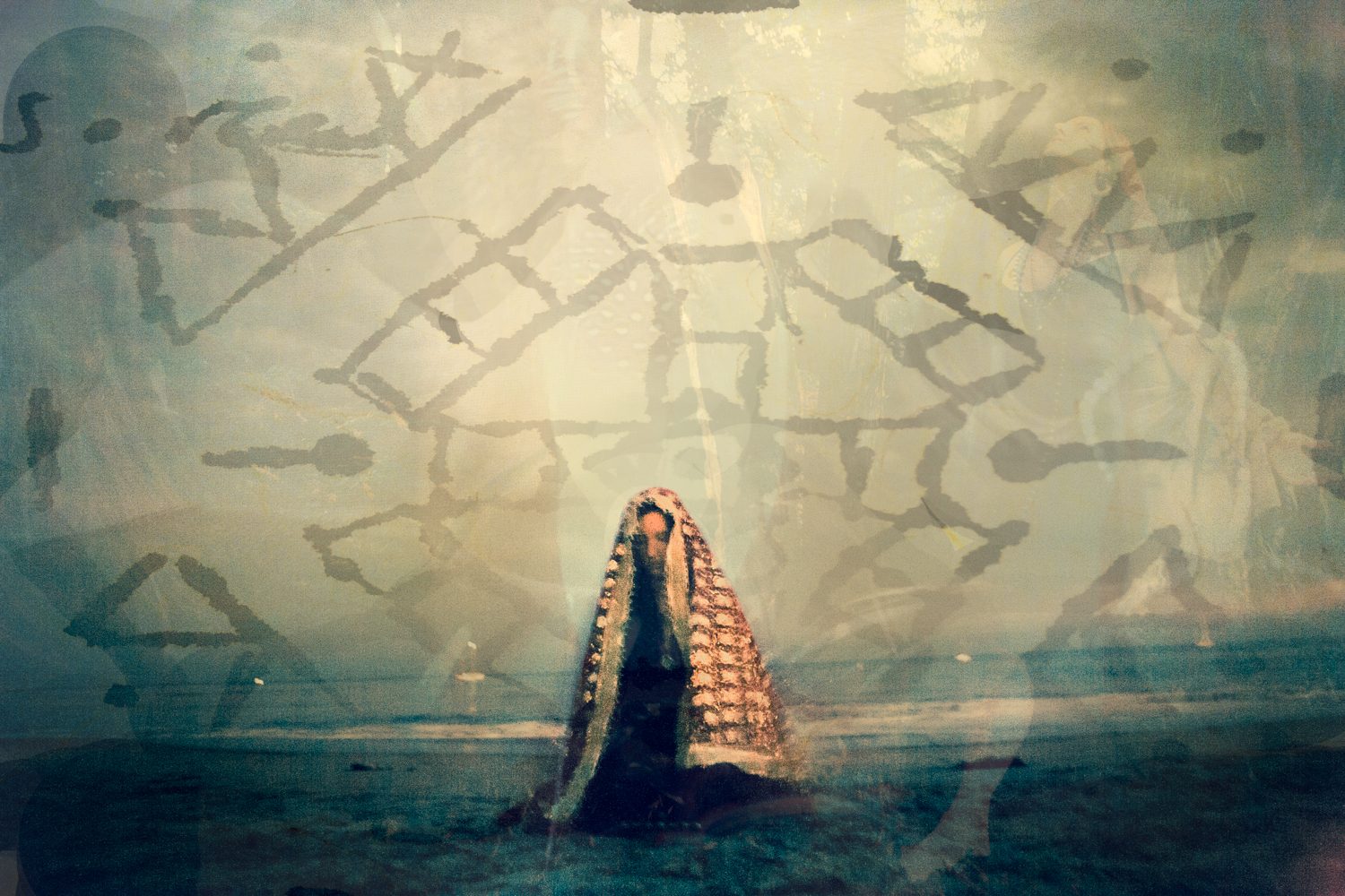 Photograph by Heather Brand: a veiled woman in a patterned shawl kneeling on a shoreline beneath layered painted symbols.