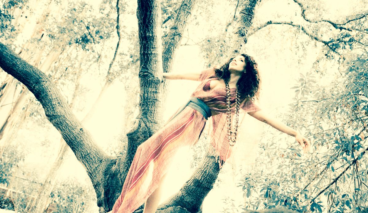 Photograph by Heather Brand: a woman leans back through the branches of an old tree, arms open and face lifted, a sheer striped dress and long beads falling loose.