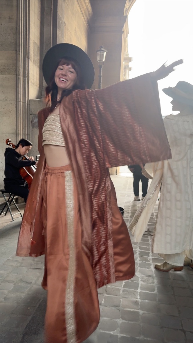 A woman in a copper silk kimono and wide hat laughs with her arms flung open beneath a Paris stone arcade, a cellist playing behind her.