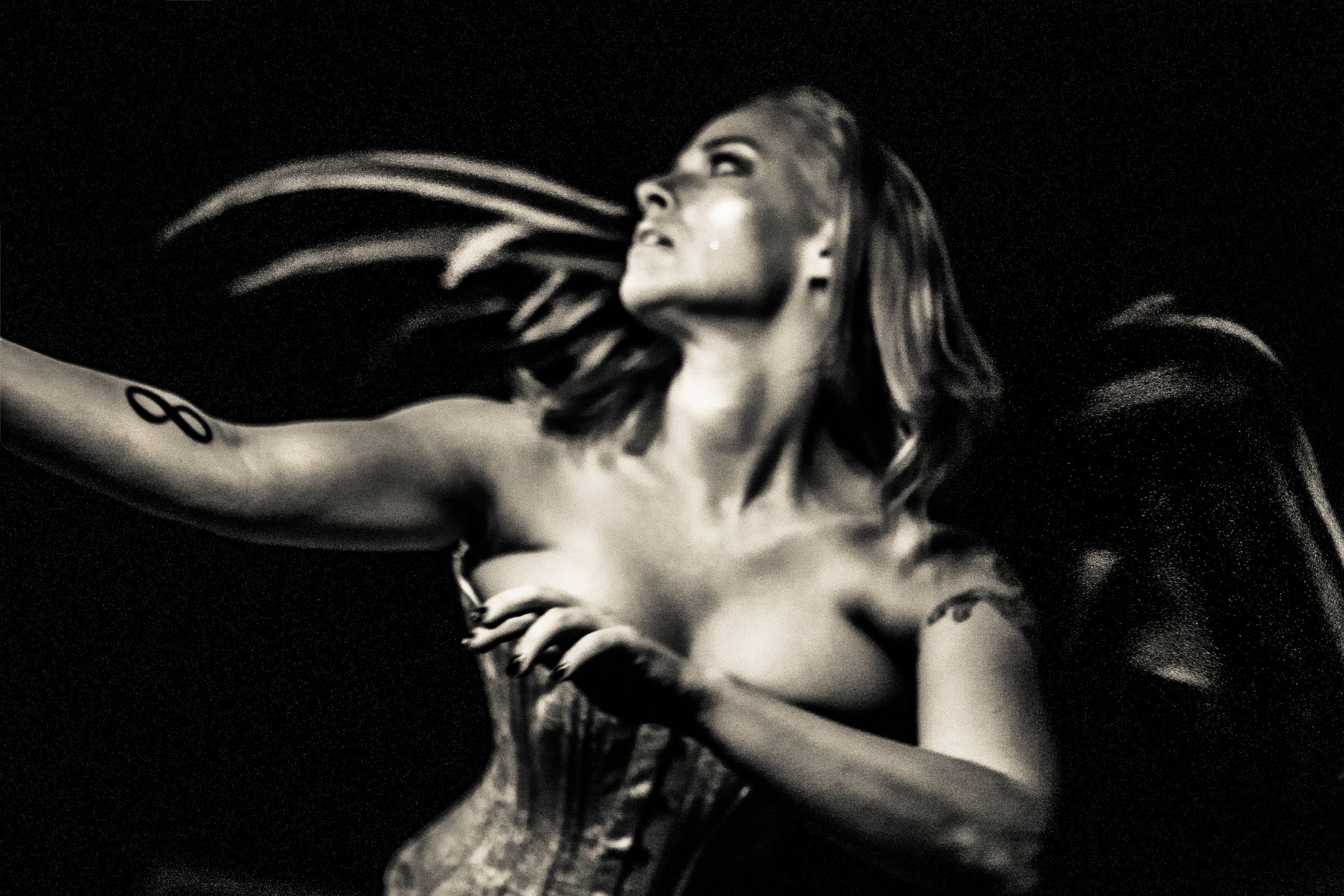 Black and white photograph by Heather Brand: a woman in darkness with her face lifted, eyes half closed, one arm extended, a spray of dark feathers sweeping behind her.