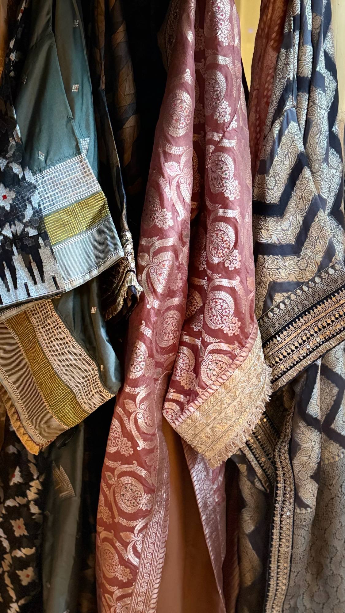 Shloaka silks hanging together on a rail: a plum brocade robe edged in gold at the centre, between teal ikat and indigo woven with gold.