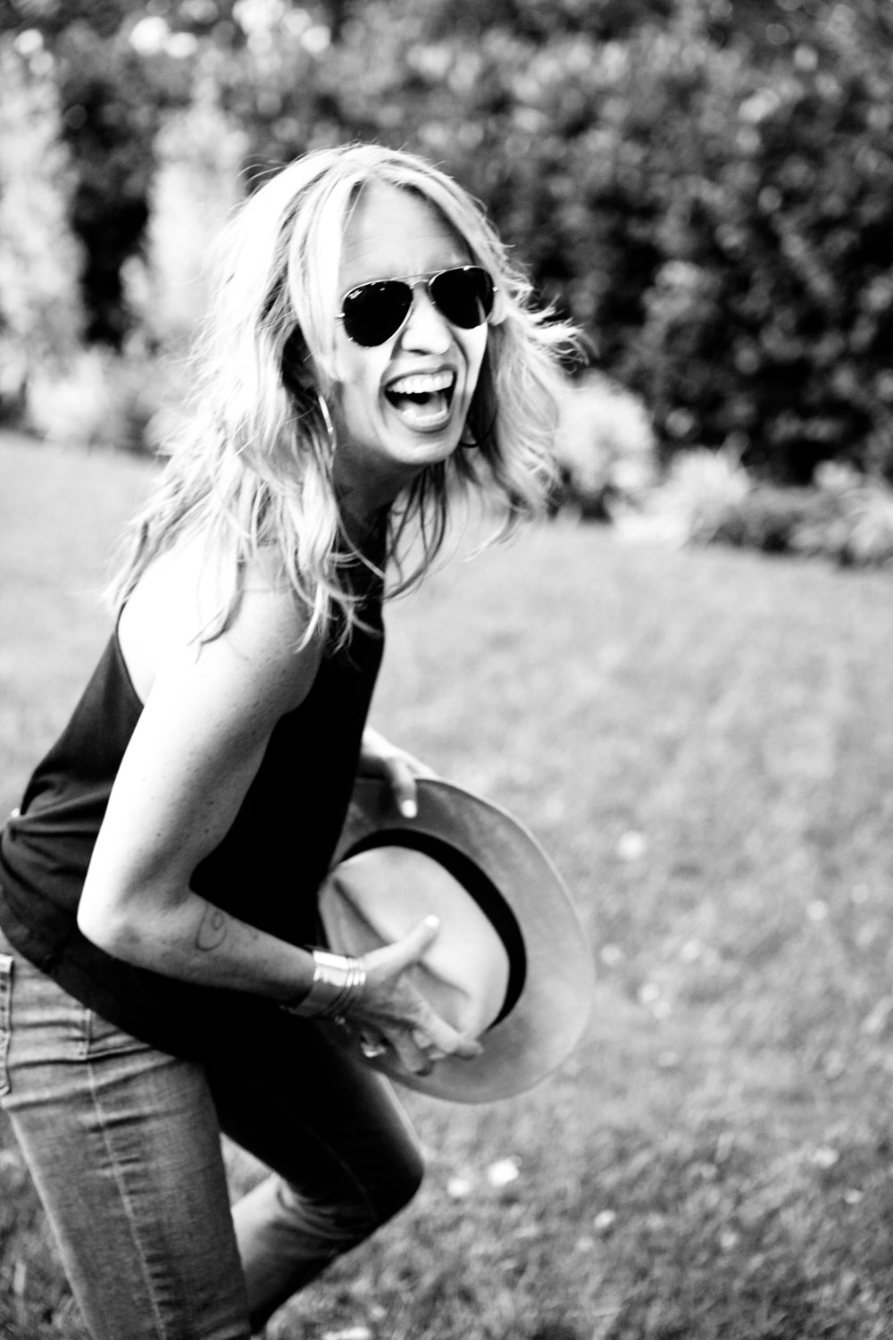 Black and white photograph by Heather Brand: a woman in sunglasses laughs out loud on a garden lawn, holding a wide brimmed hat in both hands.