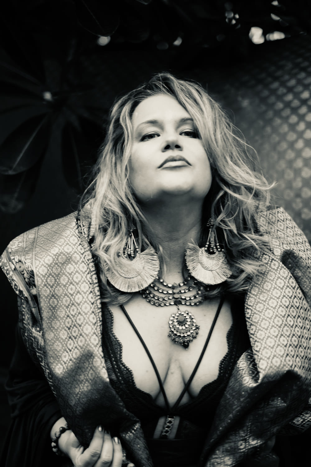 Black and white portrait of Heather Brand: chin lifted, looking straight into the lens, in a patterned brocade wrap with large fan earrings and layered necklaces.