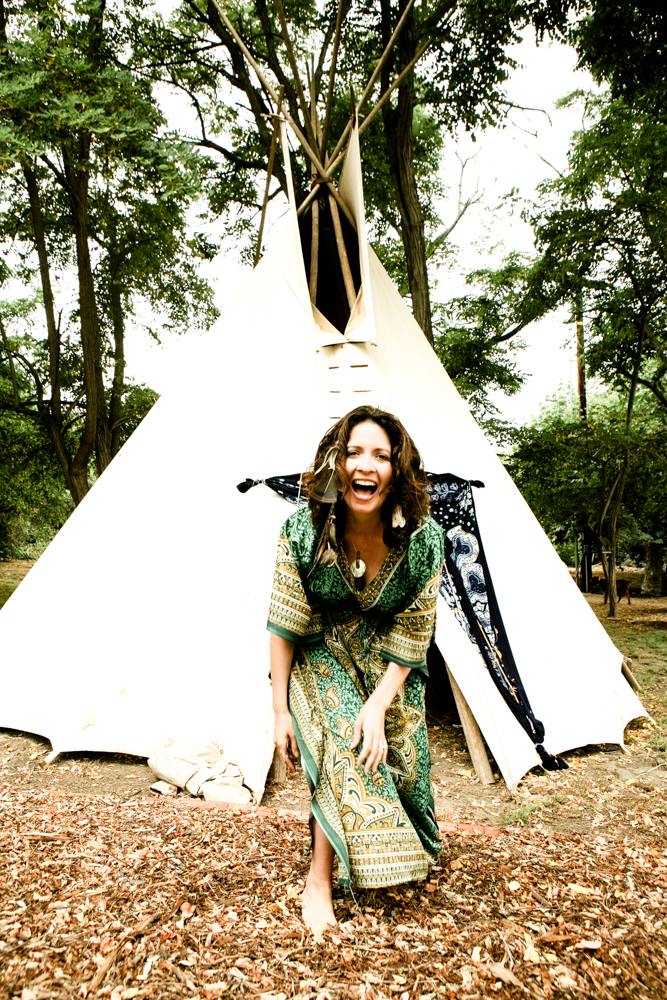 Photograph by Heather Brand: a barefoot woman in a green patterned kaftan laughs openly in front of a white teepee among tall trees.